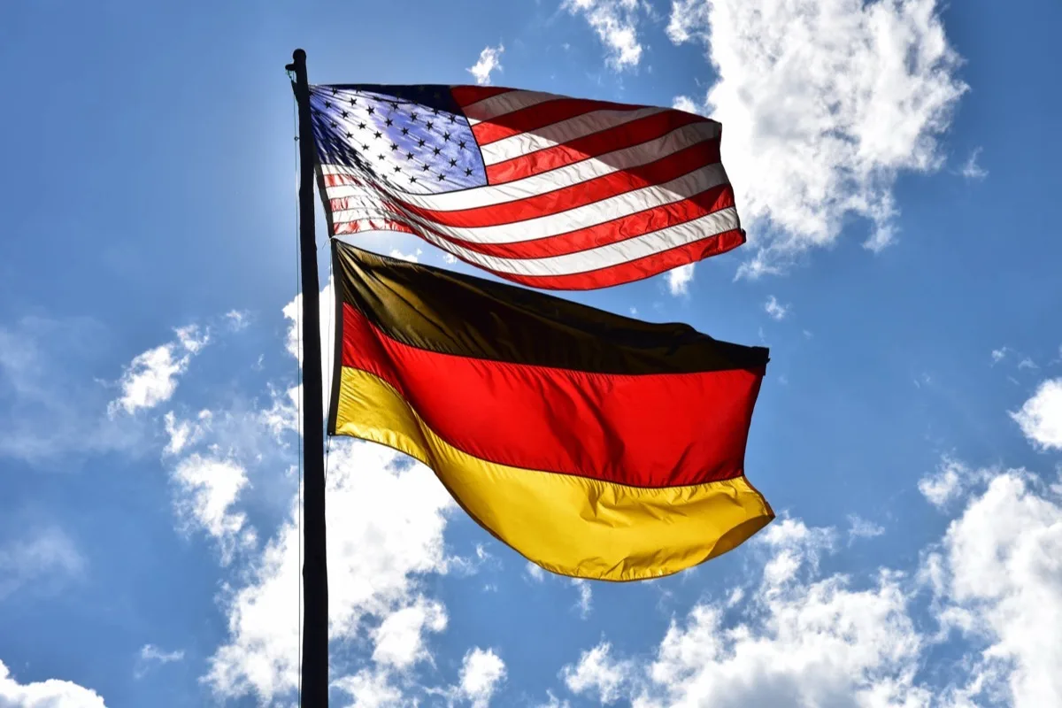 Die amerikanische und die deutsche Flagge wehen übereinander an einem Mast vor blauem Himmel.