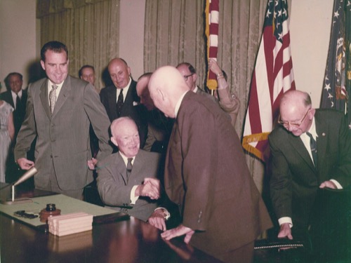 Eisenhower sitzt am Tisch der Zeremonie und gibt einem Mann die Hand, dahinter Umstehende, rechts Flaggen
